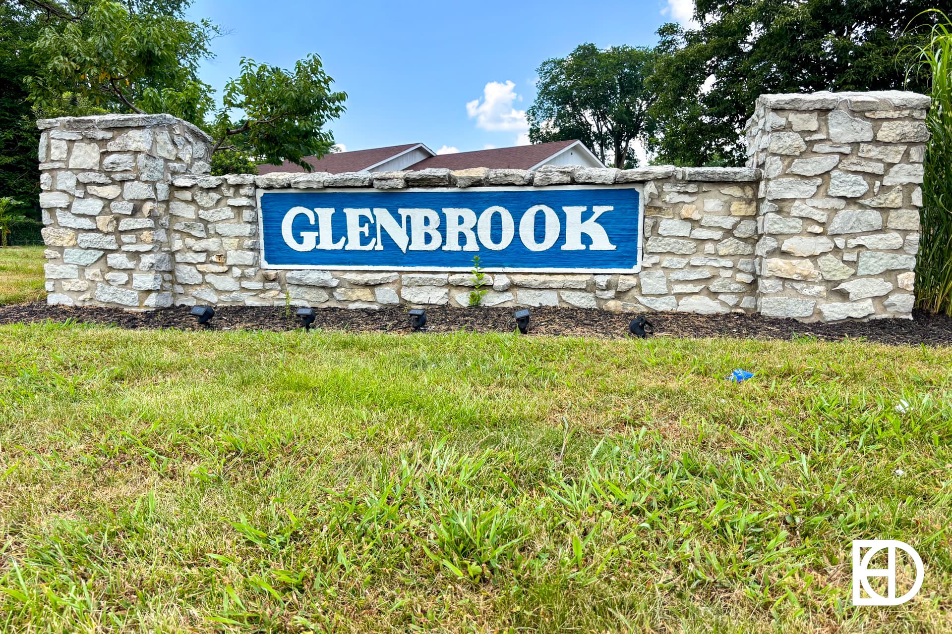 A stone sign with a blue plaque displaying the word “Glenbrook” in white capital letters, surrounded by grass, shrubs, and trees under a partly cloudy sky.
