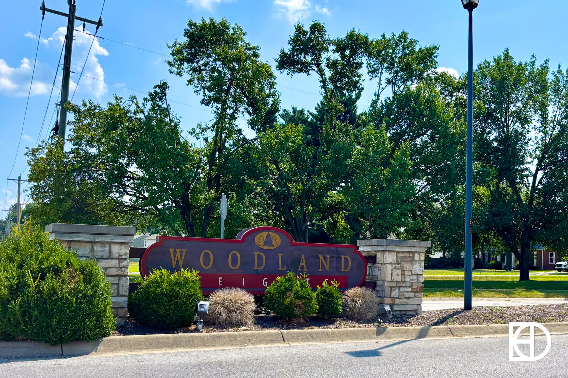 A large red and gold sign reading Woodland Heights is set in front of stone pillars and landscaping, with green trees and a grassy area visible in the background under a blue sky.