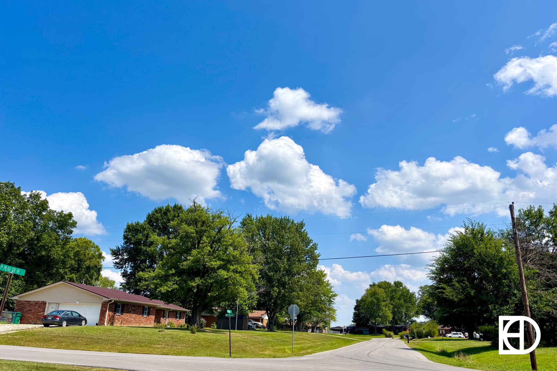 A suburban street curves past houses and green lawns under a bright blue sky with scattered white clouds. Tall trees line the road, and a basketball hoop is visible near a driveway.