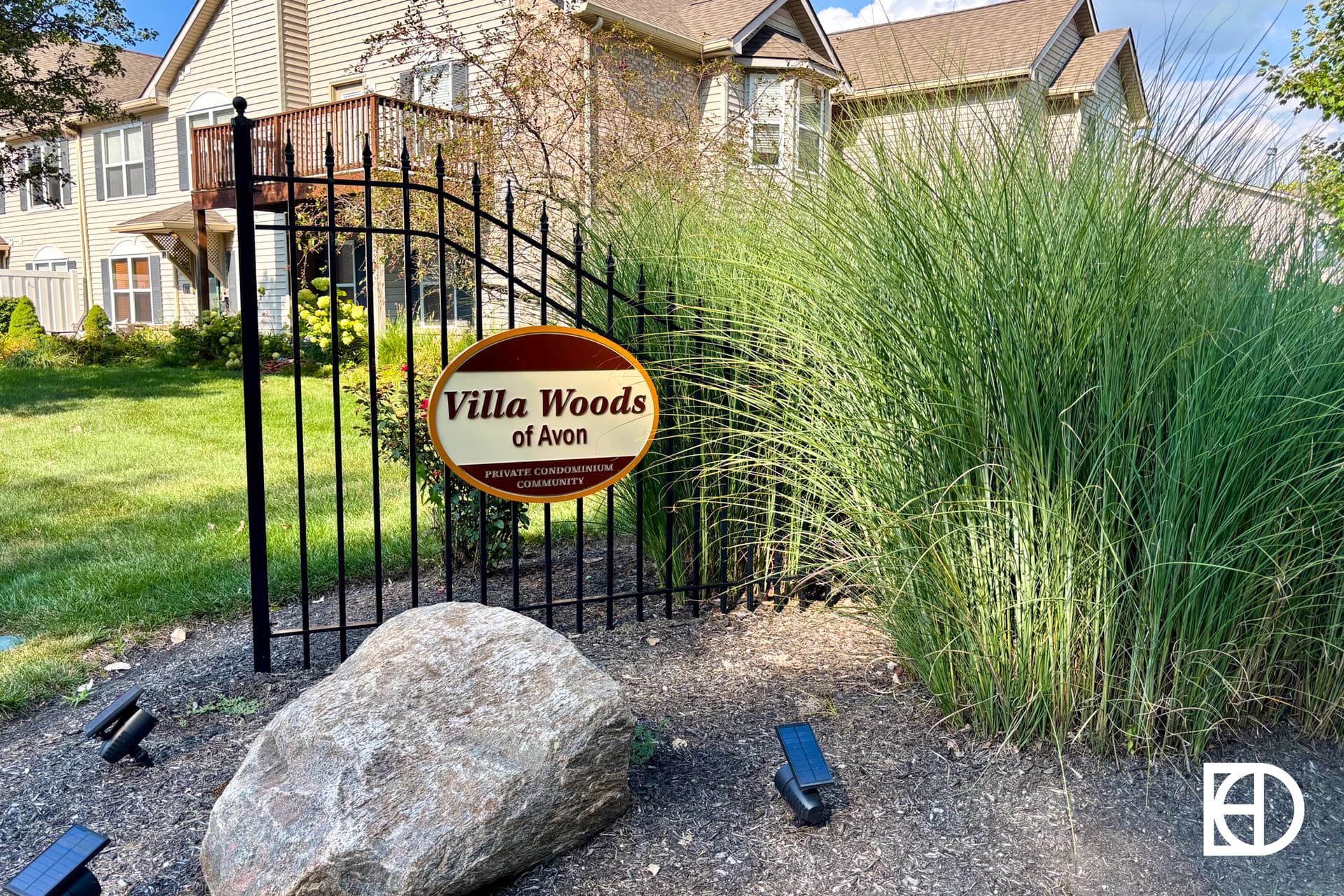 A black metal gate with a sign reading “Villa Woods of Avon” stands in front of tall ornamental grasses and suburban houses on a sunny day; a large rock and outdoor lights decorate the foreground.