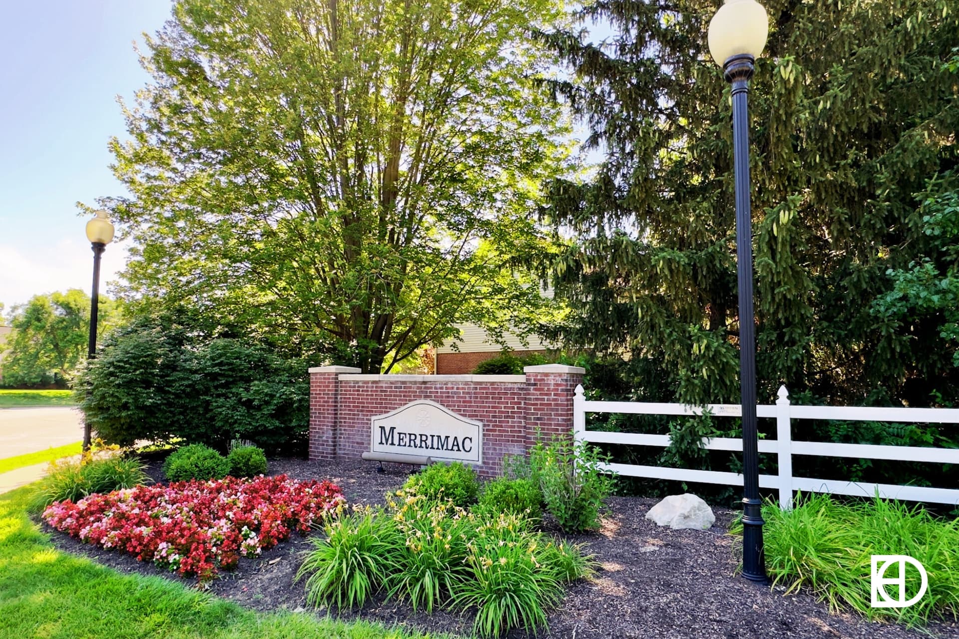A brick sign reading Merrimac sits near a white fence, lamp posts, flowerbeds, and trees in a landscaped area.