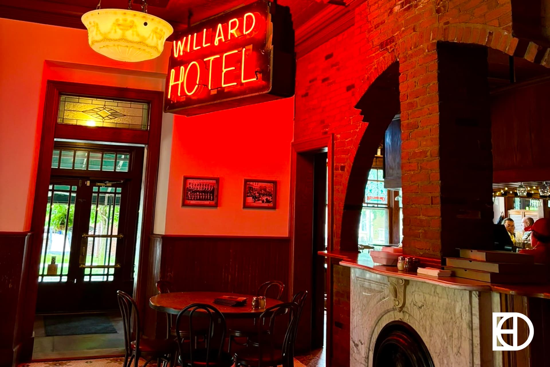 A warmly lit vintage hotel lobby with red neon Willard Hotel sign, wooden furniture, exposed brick, a stained glass door, and a marble fireplace.