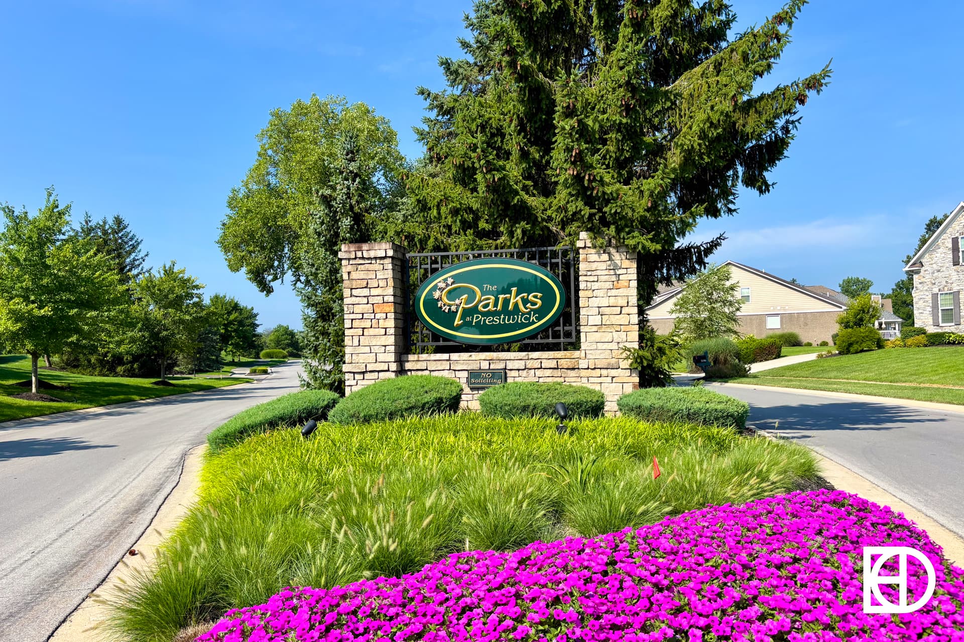 A stone sign reads Parks at Prestwick, surrounded by neatly trimmed bushes, green grass, vibrant purple flowers, and residential houses on a sunny day.