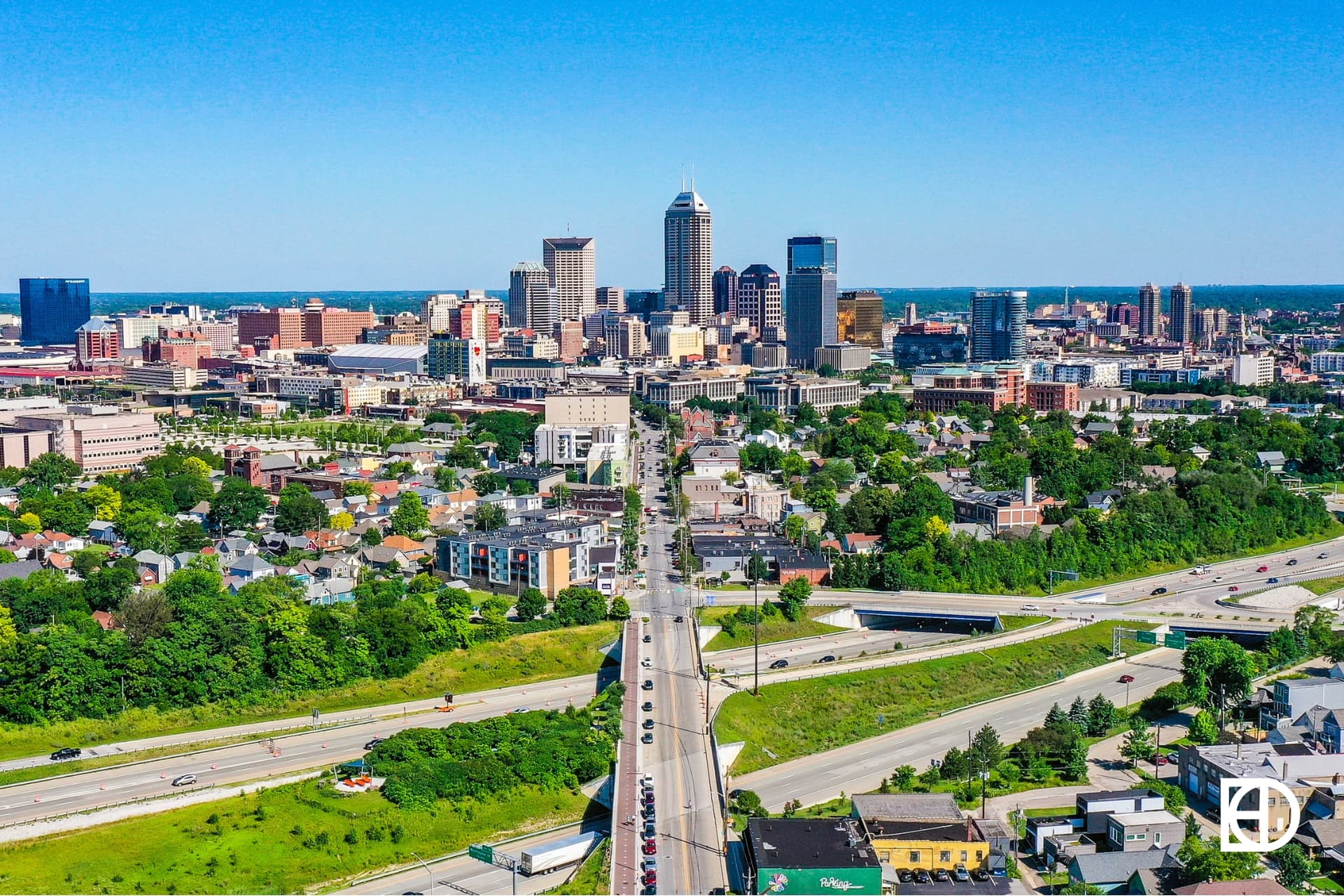 Aerial view of downtown Indianapolis with city skyscrapers, residential areas, and highways under a clear blue sky.