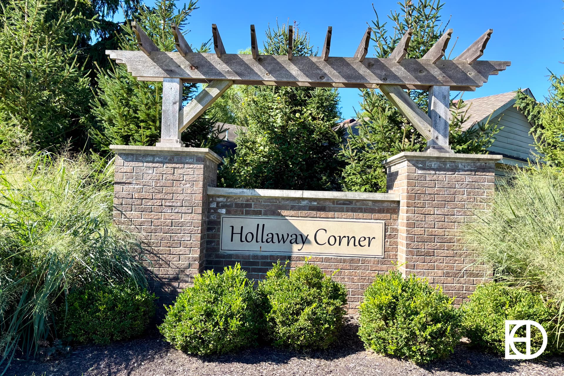 A brick entrance sign reading Hollaway Corner, surrounded by green bushes, tall grasses, and evergreen trees, with a wooden pergola structure above the sign on a sunny day.
