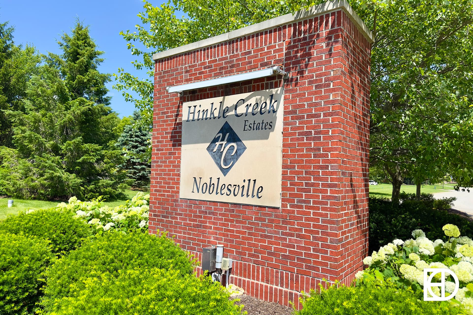 A brick entrance sign for Hinkle Creek Estates in Noblesville, surrounded by green bushes, white flowers, and trees on a sunny day.