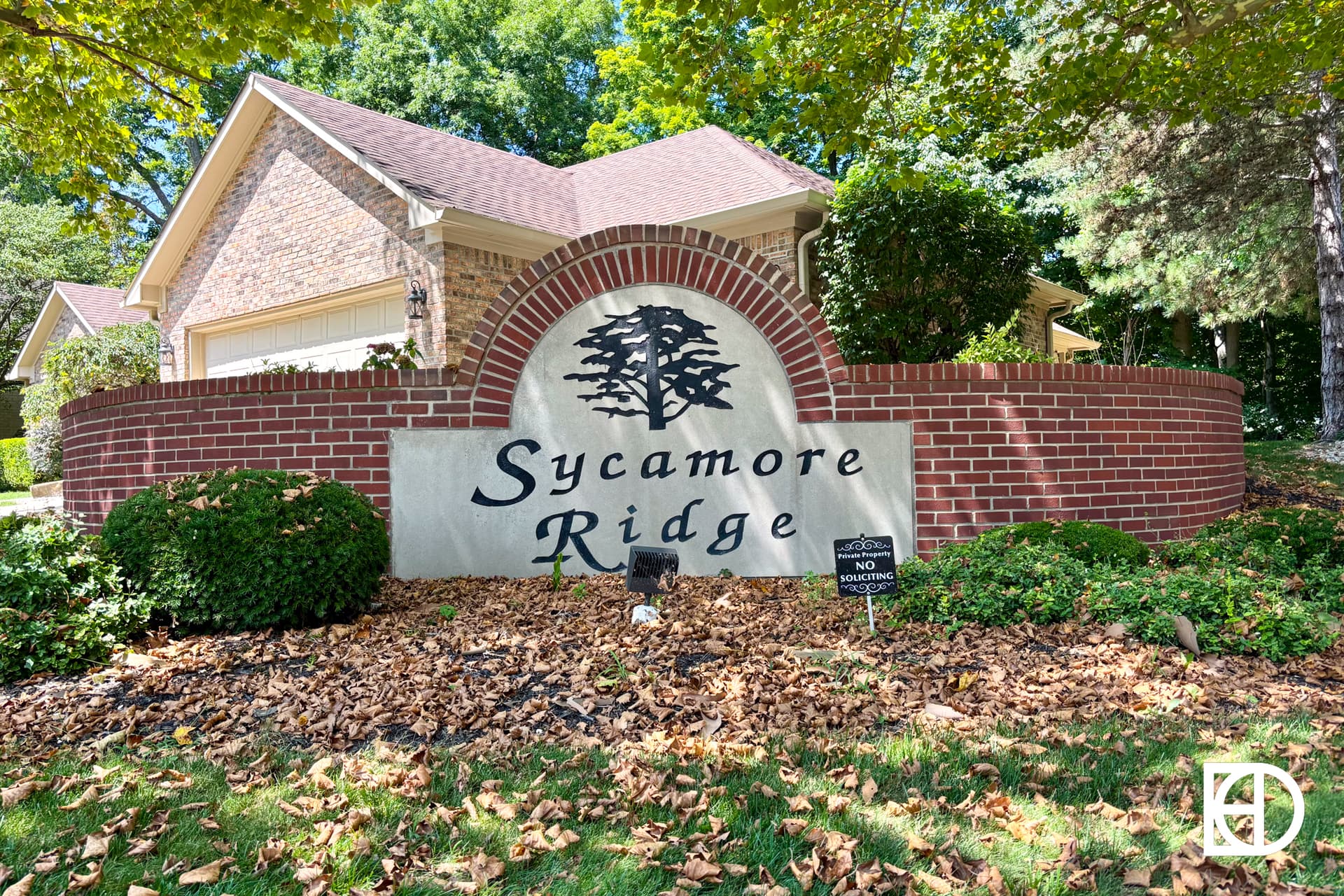 A brick sign with Sycamore Ridge and a tree logo stands in front of a house, surrounded by green grass and fallen leaves. A small sign in front reads No Soliciting. Trees and shrubs are in the background.