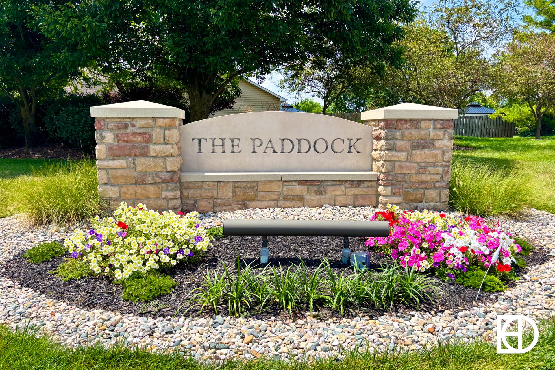 A landscaped stone sign reads THE PADDOCK, surrounded by colorful flowers, green grass, and small decorative rocks, with trees and a house in the background. A bench is placed in front of the sign.