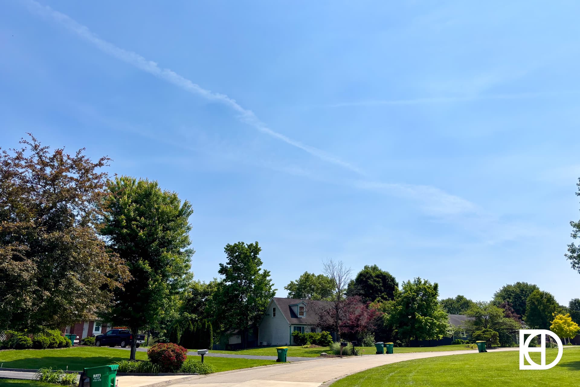 A suburban neighborhood street with green lawns, trees, houses, and several green garbage bins on a sunny day under a blue sky.