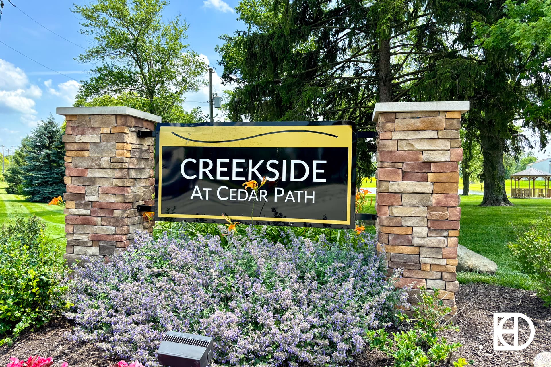 A stone sign surrounded by purple flowers and greenery reads Creekside at Cedar Path with a yellow wave design, set against trees and a partly cloudy sky.