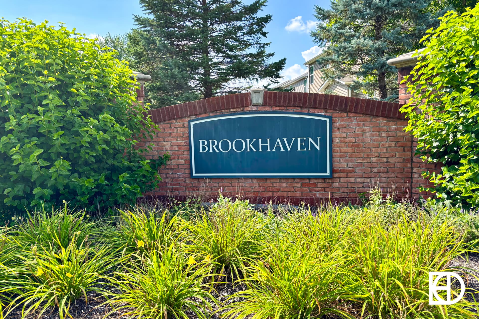 A brick entrance sign reading BROOKHAVEN is surrounded by green bushes and grass, with trees and a sunny blue sky in the background. A white logo is in the bottom right corner.