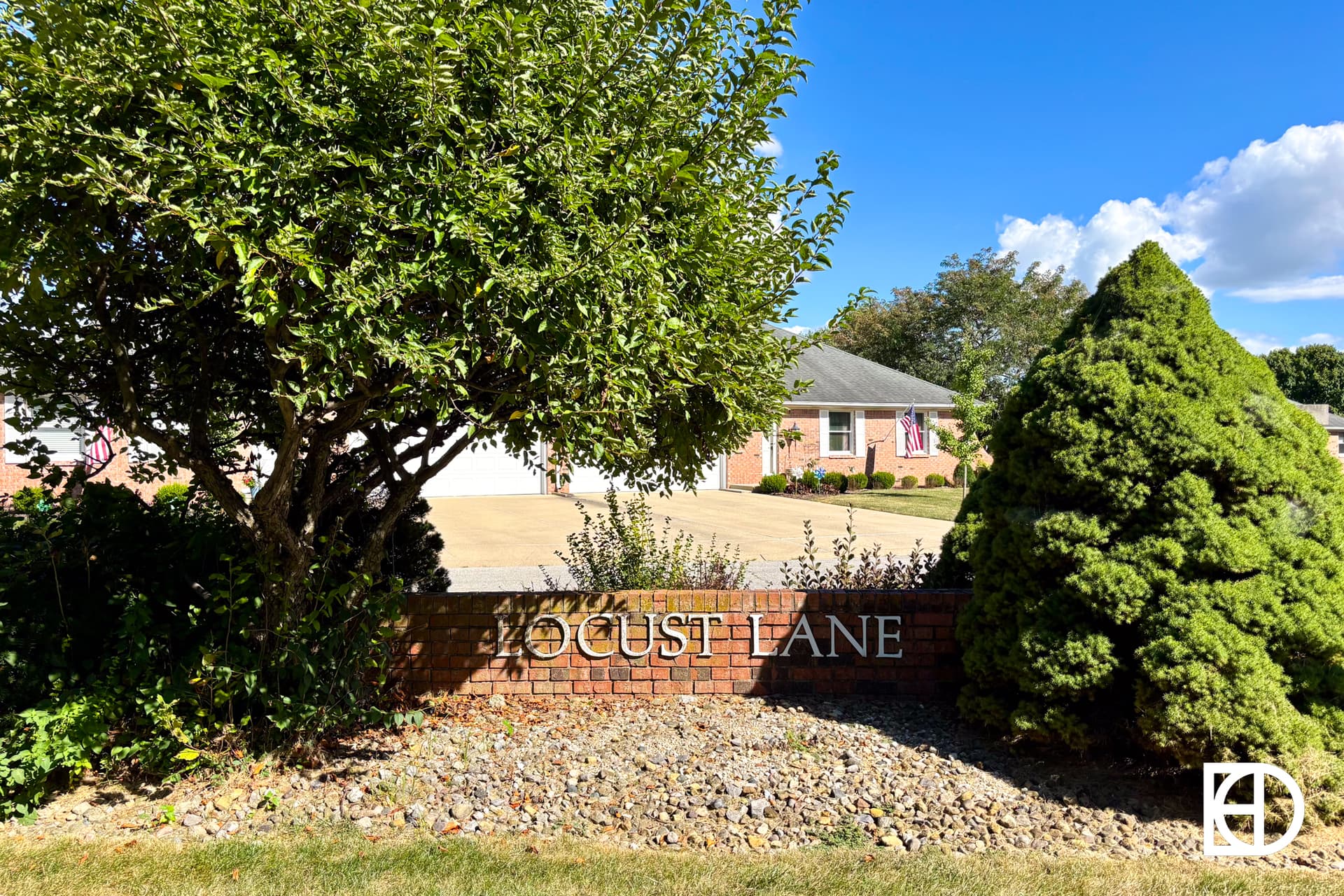 A brick sign reading LOCUST LANE stands in front of a suburban house, surrounded by neatly trimmed trees, shrubs, and landscaping rocks under a blue sky with scattered clouds.
