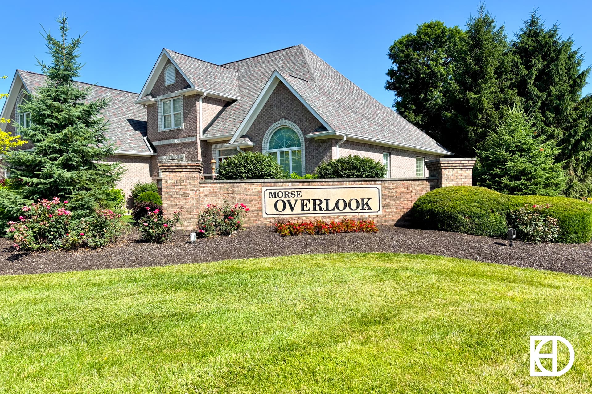 A brick entrance sign reads Morse Overlook in front of a landscaped lawn with bushes, flowers, and a large house with a peaked roof in the background. A white logo is in the bottom right corner.