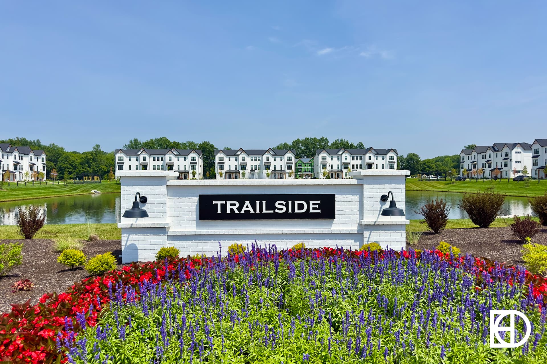 A landscaped garden with red and purple flowers surrounds a white brick sign reading TRAILSIDE. Behind the sign is a pond and rows of white townhouses under a clear blue sky.