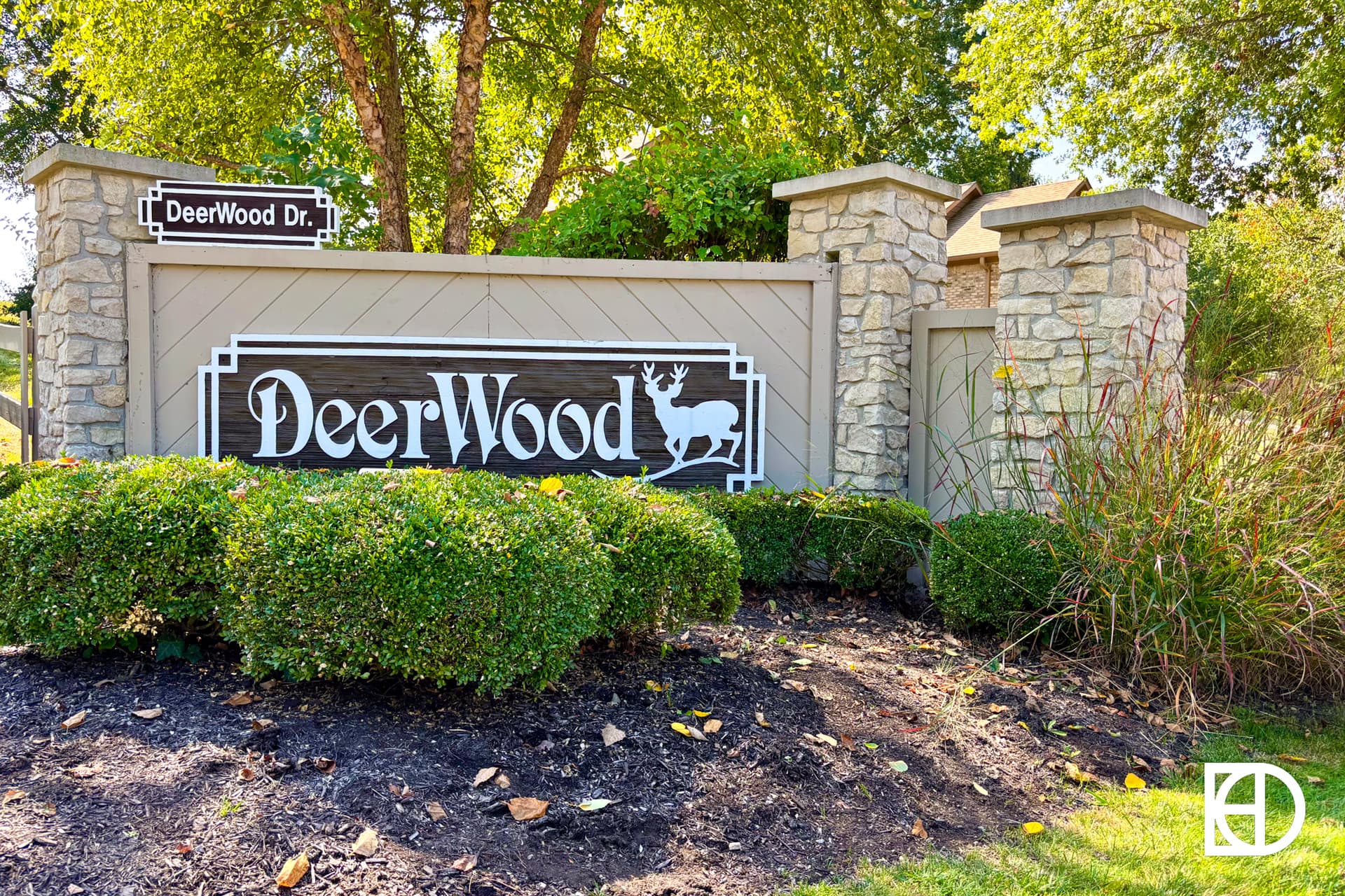 A suburban neighborhood street with modern houses, green lawns, and parked cars under a bright blue sky filled with fluffy white clouds.