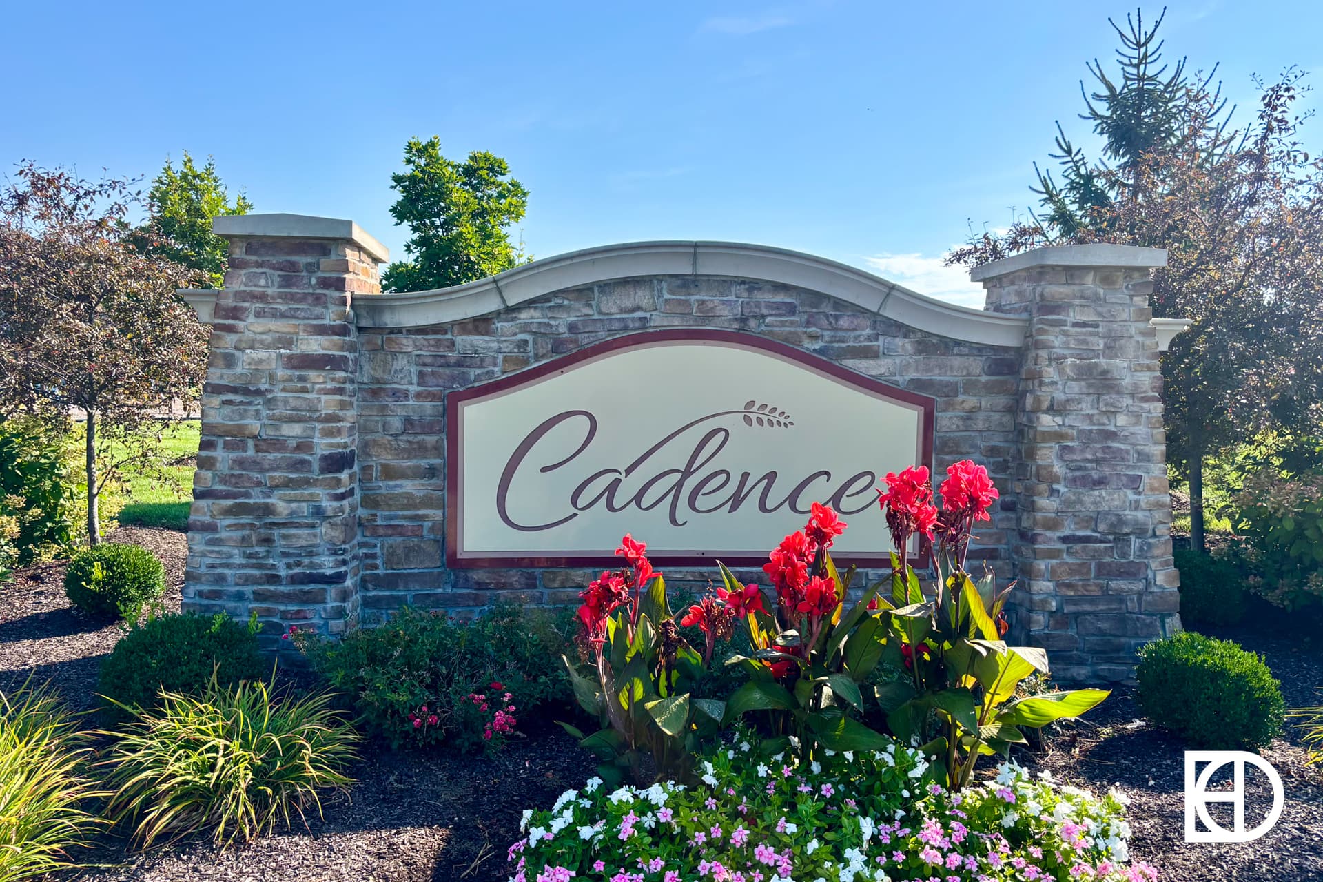 A stone entrance sign reading Cadence, framed by landscaped bushes and red flowers, stands under a clear blue sky with trees in the background.