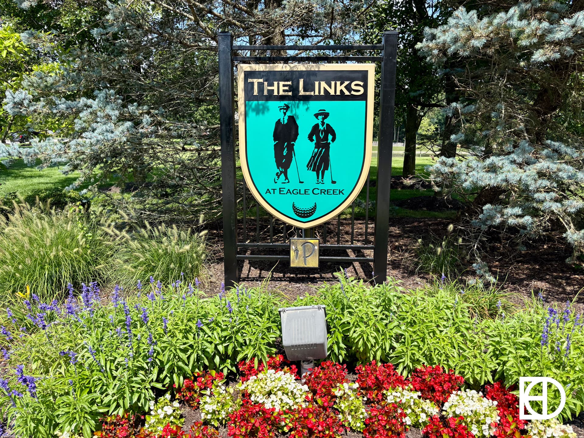 A sign reading The Links at Eagle Creek stands among colorful flowers and greenery, featuring silhouettes of two golfers and golf clubs against a green background.