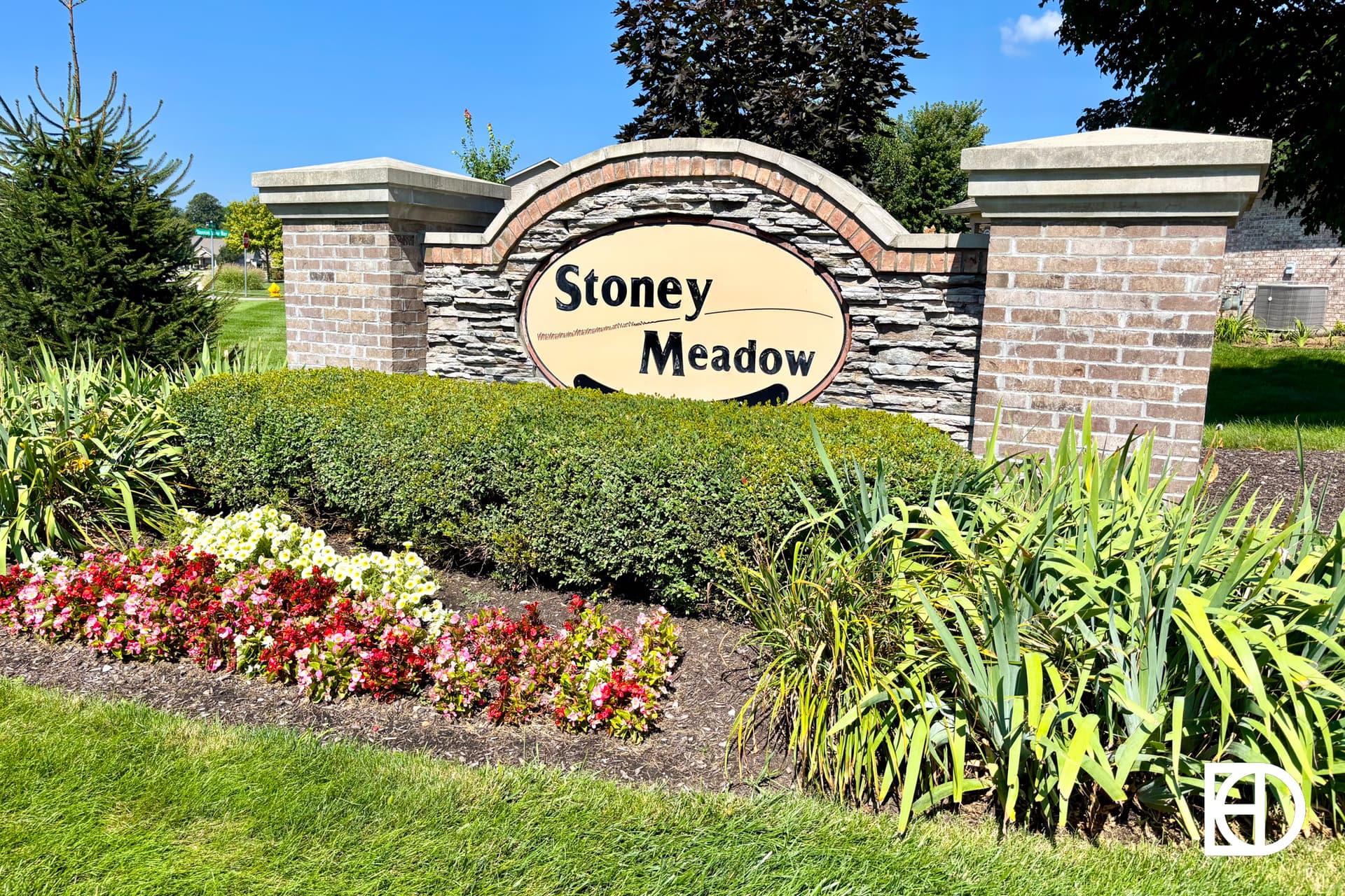 A brick and stone entrance sign reads Stoney Meadow, surrounded by green shrubs, colorful flowers, and landscaping on a sunny day.