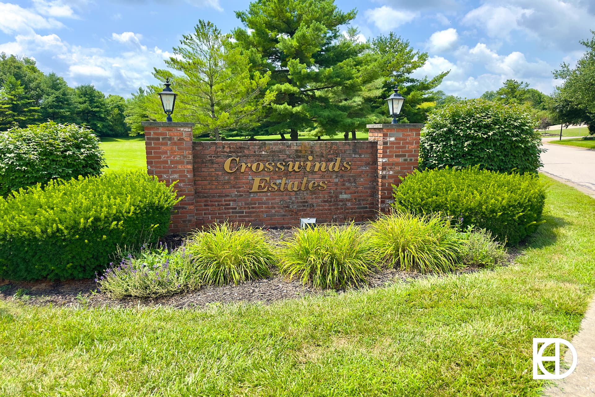 Brick sign reading Crosswinds Estates surrounded by green bushes and trees, with two lanterns on top of the sign, set against a grassy area and blue sky with clouds.
