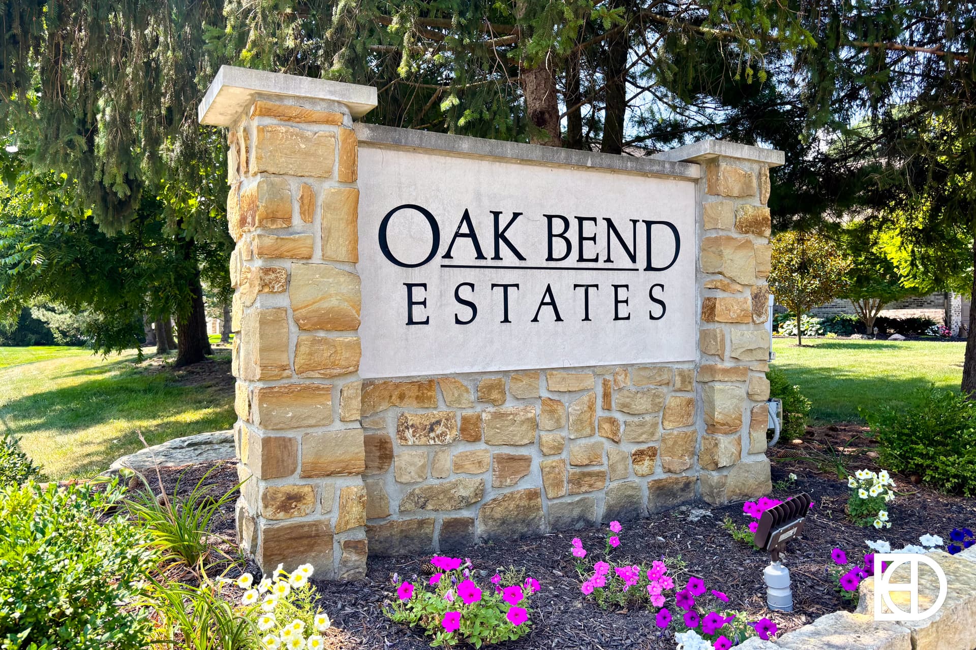 A stone sign reads OAK BEND ESTATES in black letters, surrounded by green trees, grass, and colorful flowers at the base. The area looks well-maintained and inviting.