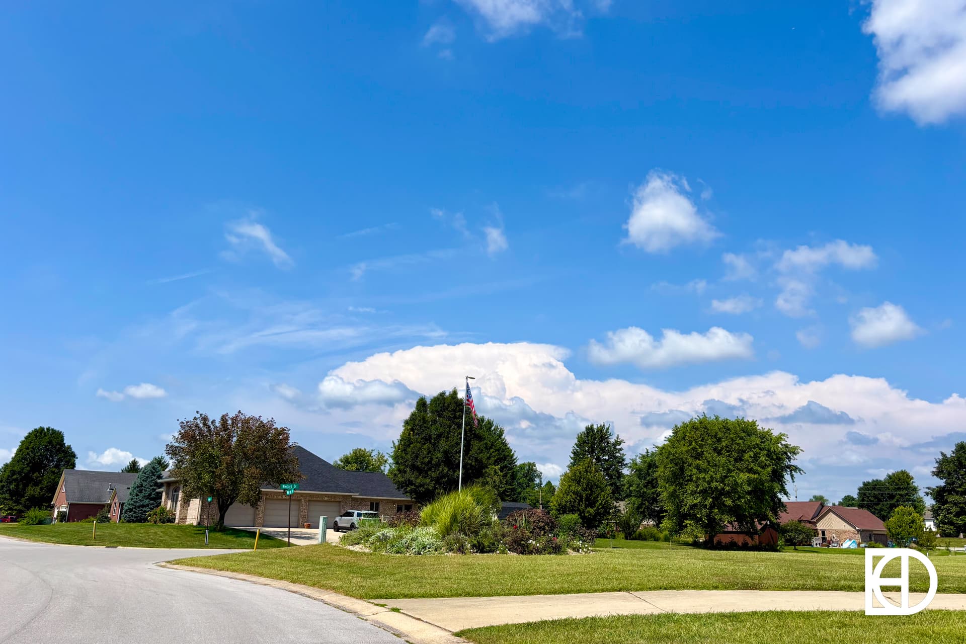 A suburban neighborhood with houses, green lawns, trees, and a clear blue sky with scattered white clouds. A street curves through the scene, and a logo is visible in the bottom right corner.