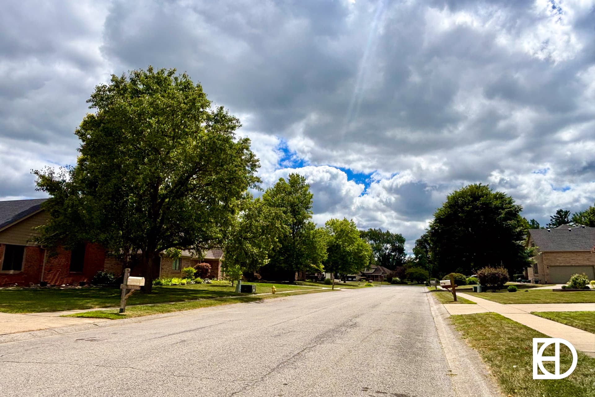 A quiet suburban street with houses, green lawns, and trees under a partly cloudy sky. The scene is peaceful with no people or cars visible. A white logo appears in the bottom right corner.