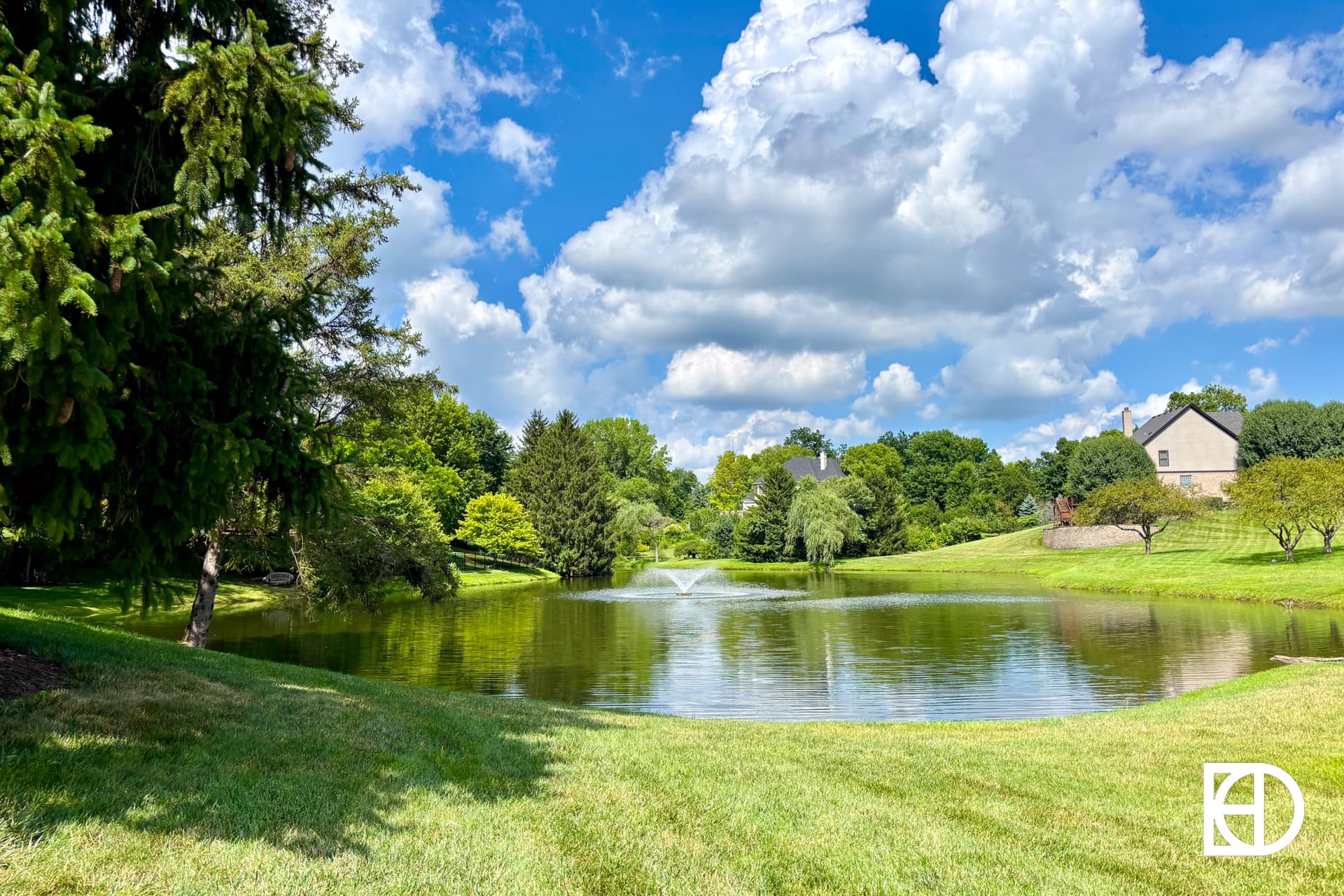 A small pond with a fountain surrounded by green grass and trees, under a blue sky with scattered clouds. Two houses are visible in the background on a slightly elevated hill.