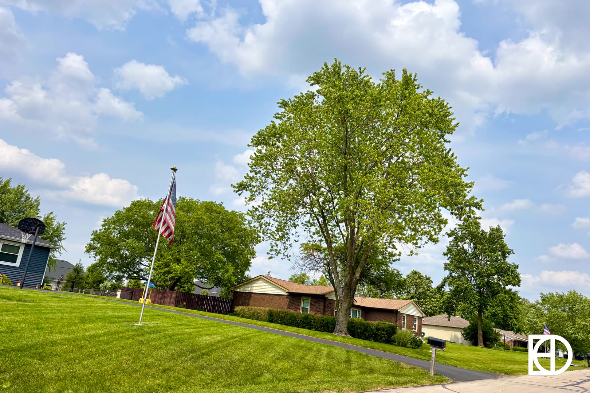 A single-story brick house with a green lawn, a large tree in front, and an American flag on a pole. The sky is partly cloudy.