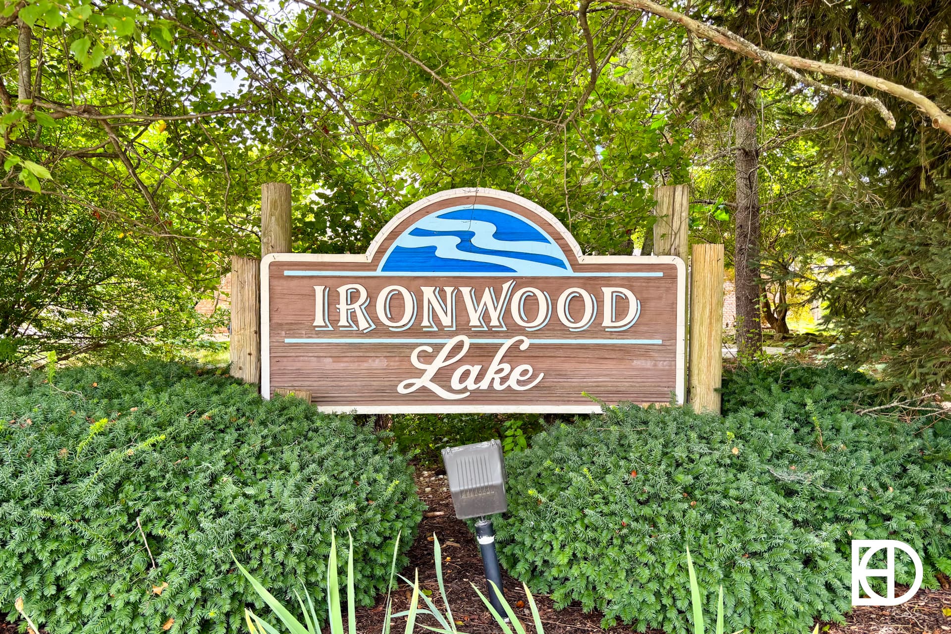 A wooden sign reading Ironwood Lake with a blue wave design sits among green bushes and trees, with a small light fixture in front.