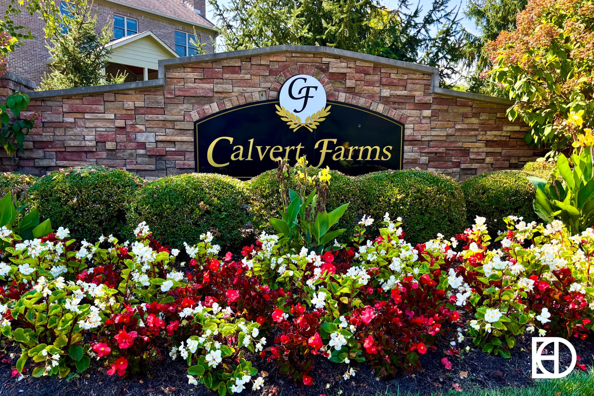 A stone sign reads Calvert Farms with CF above it, surrounded by manicured bushes and colorful red and white flowers. A large house and tall trees are visible in the background.