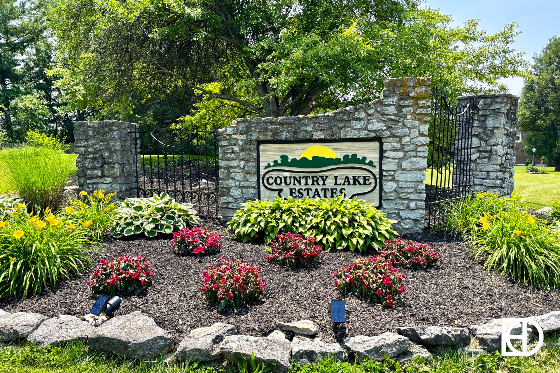 Stone entrance sign for Country Lake Estates surrounded by colorful flowers, green plants, and mulch, with a backdrop of grass, trees, and a sunny blue sky.