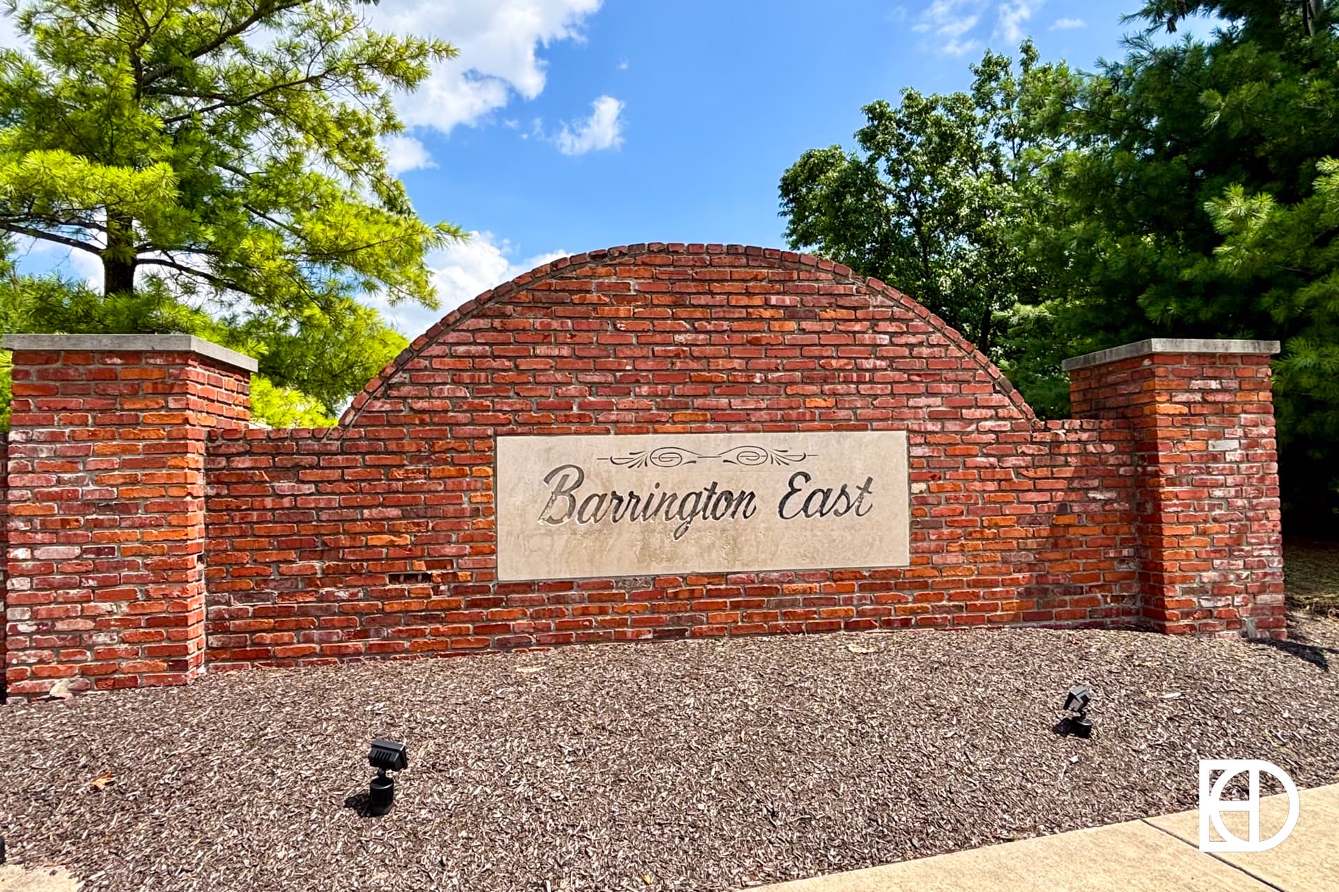 A brick entrance sign with a stone plaque reading Barrington East, surrounded by trees and mulch under a partly cloudy sky.