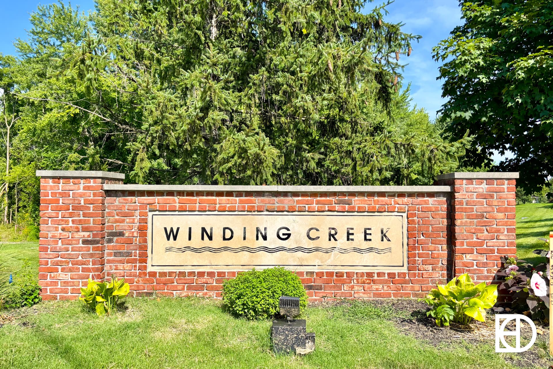 A brick sign with the words Winding Creek surrounded by green grass, small shrubs, and trees in the background on a sunny day.