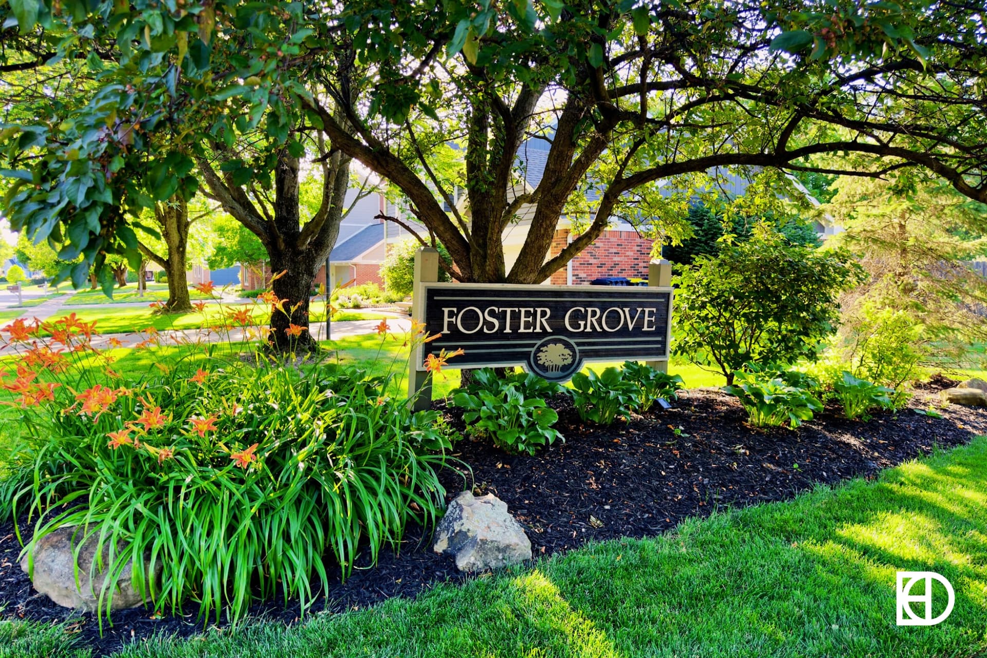 Photo of entrance sign to Foster Grove with daylilies and shrubs surrounding and trees in the background.