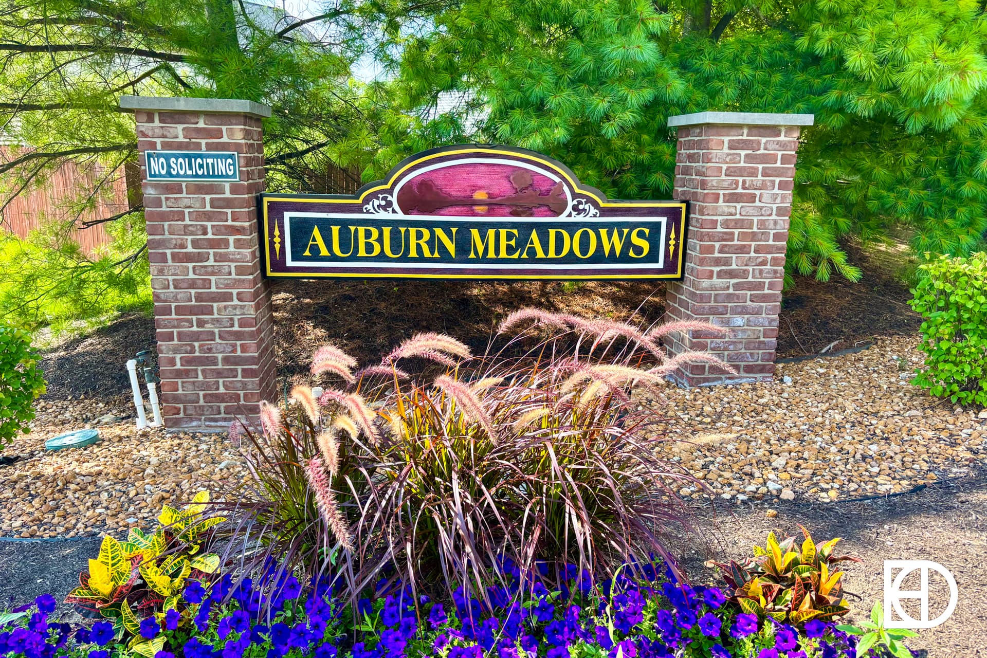 A brick sign for Auburn Meadows stands among green shrubs and ornamental grasses, with purple flowers and a No Soliciting sign on the left pillar.