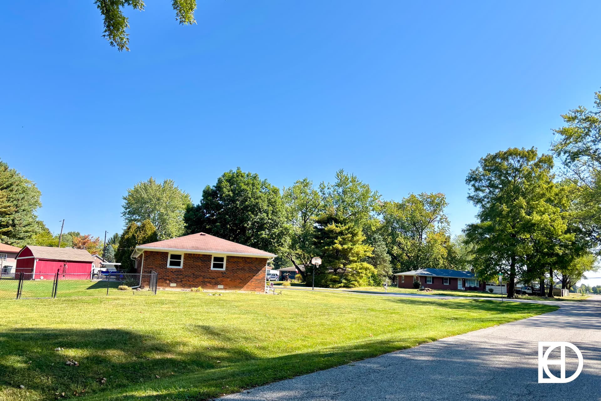 A small brick house with a red roof sits on a grassy lawn beside a quiet street, surrounded by trees under a clear blue sky. Other houses and a red barn are visible in the background.