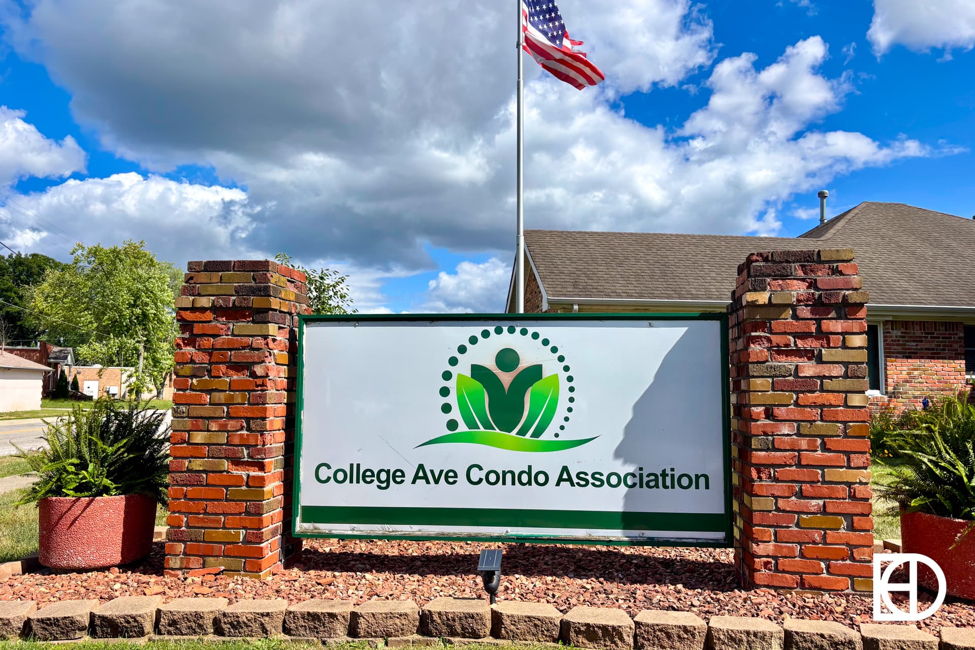 A brick sign with green and white lettering reads College Ave Condo Association. There’s a plant logo above the text, with an American flag and a house roof visible in the background under a blue sky.