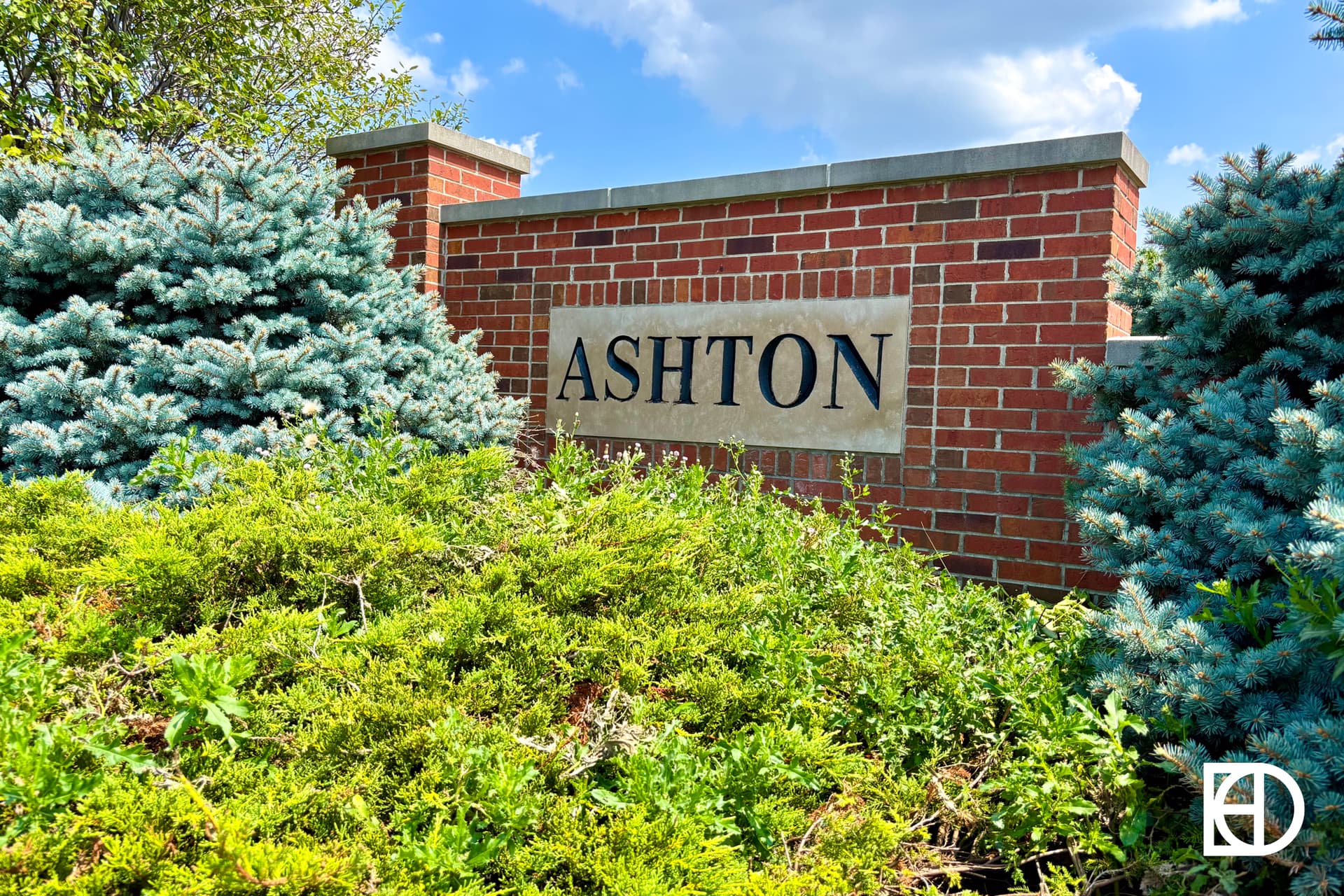 A brick entrance sign reading ASHTON is surrounded by green bushes and blue spruce trees under a partly cloudy sky.