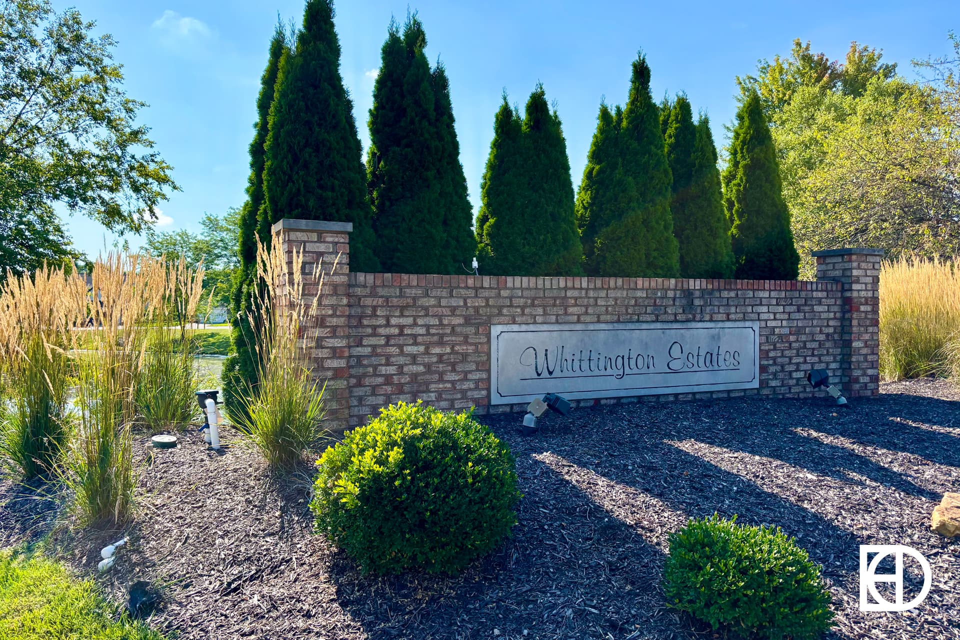 A brick entrance sign for Whittington Estates is surrounded by tall grasses, bushes, and evergreen trees under a clear blue sky.