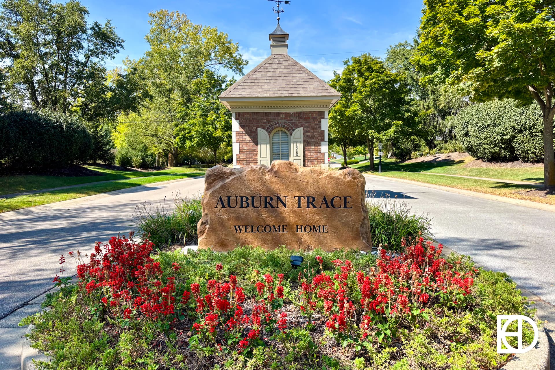 A large stone sign reading Auburn Trace Welcome Home stands surrounded by red flowers at the entrance of a residential area, with a small brick building and trees in the background under a clear blue sky.