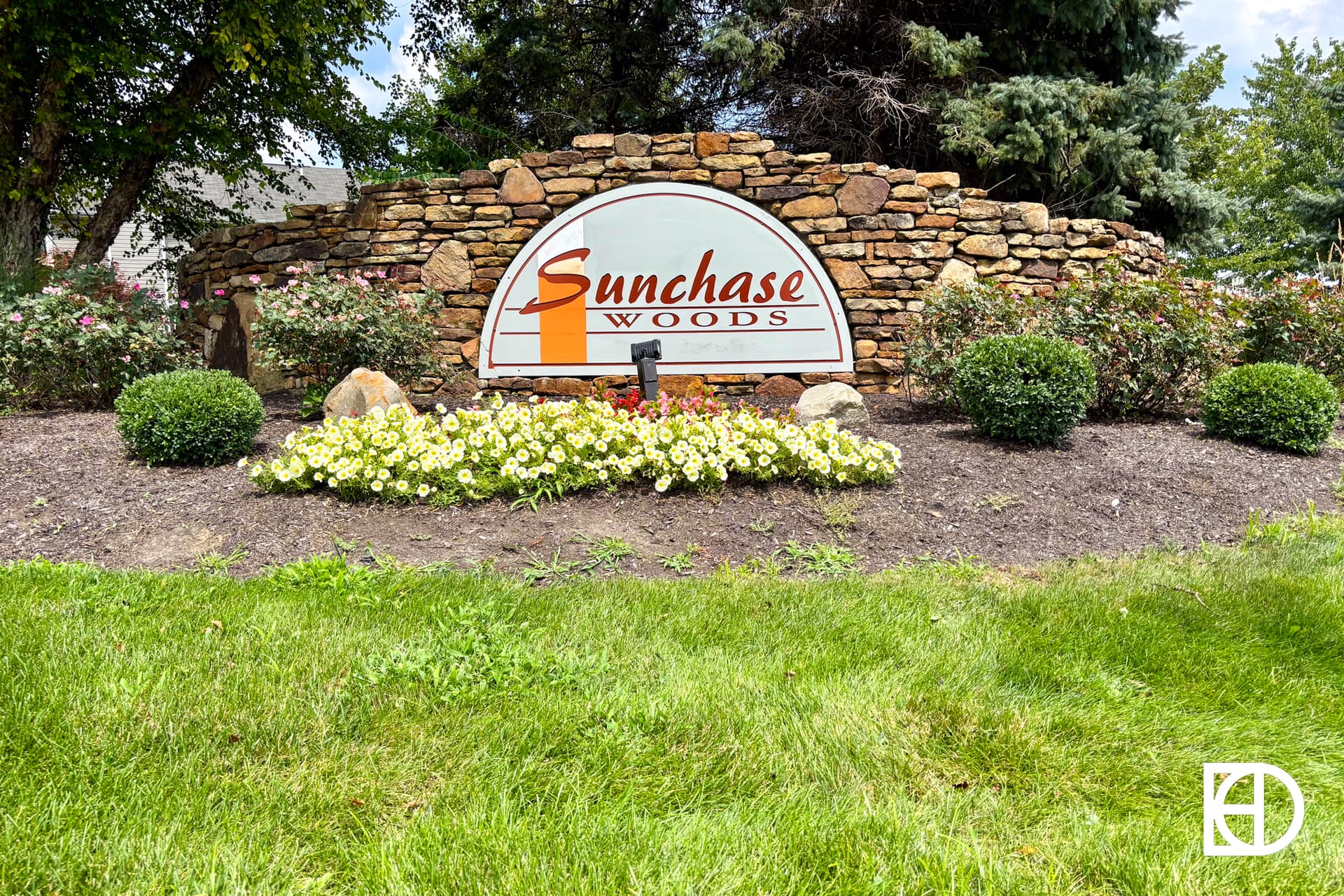 A stone sign reading Sunchase Woods is surrounded by bushes, flowers, green grass, and trees, with a blue sky in the background.