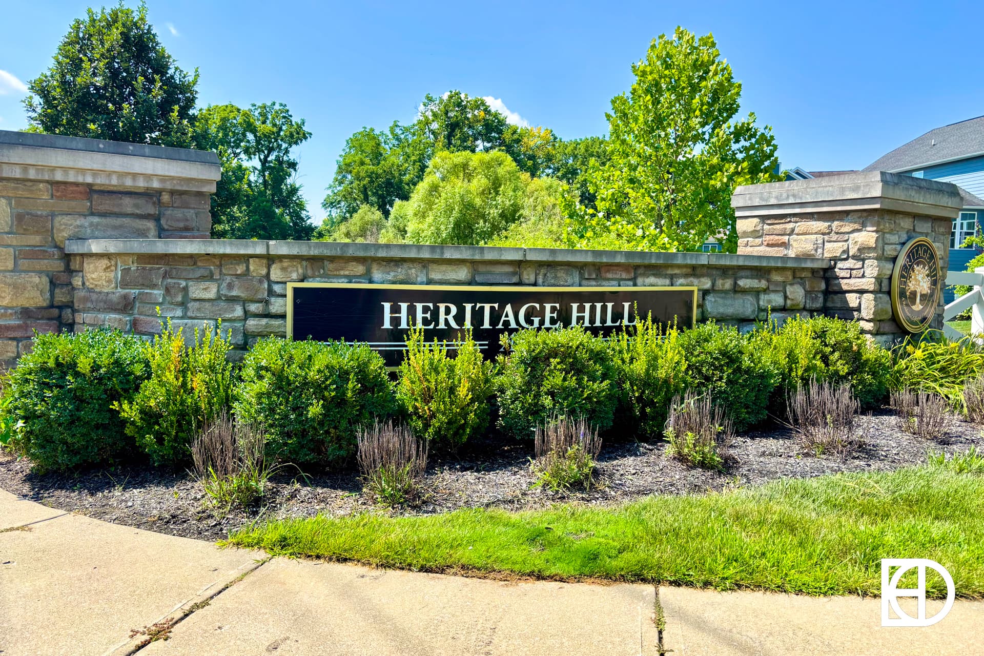 A stone sign reading HERITAGE HILL is surrounded by green bushes and landscaping, with trees and a blue sky in the background. A white logo is in the lower right corner of the image.
