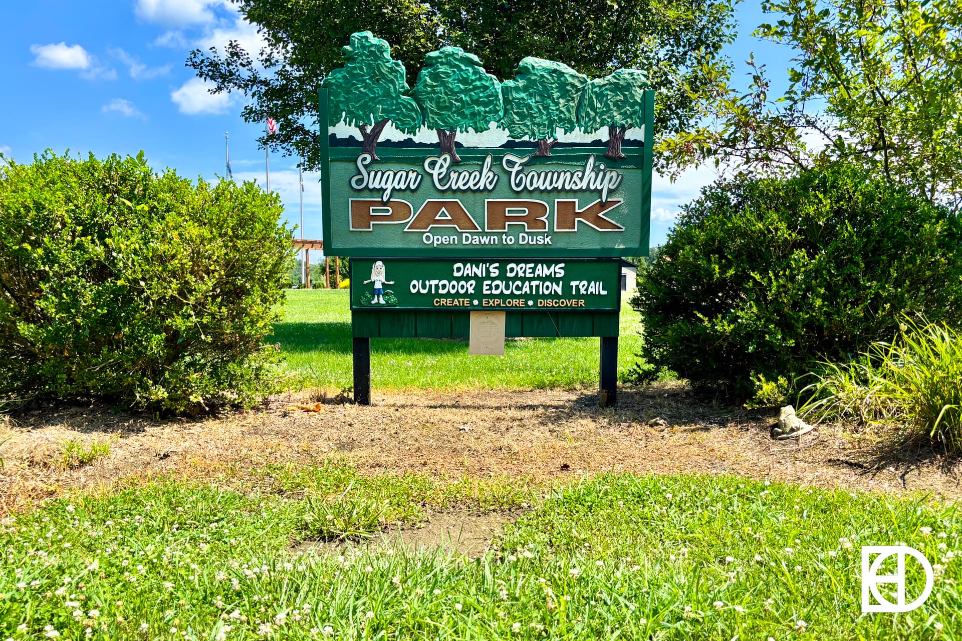 A green sign reads Sugar Creek Township Park, Open Dawn to Dusk with Danis Dreams Outdoor Education Trail below it, surrounded by bushes, grass, and trees under a blue sky.
