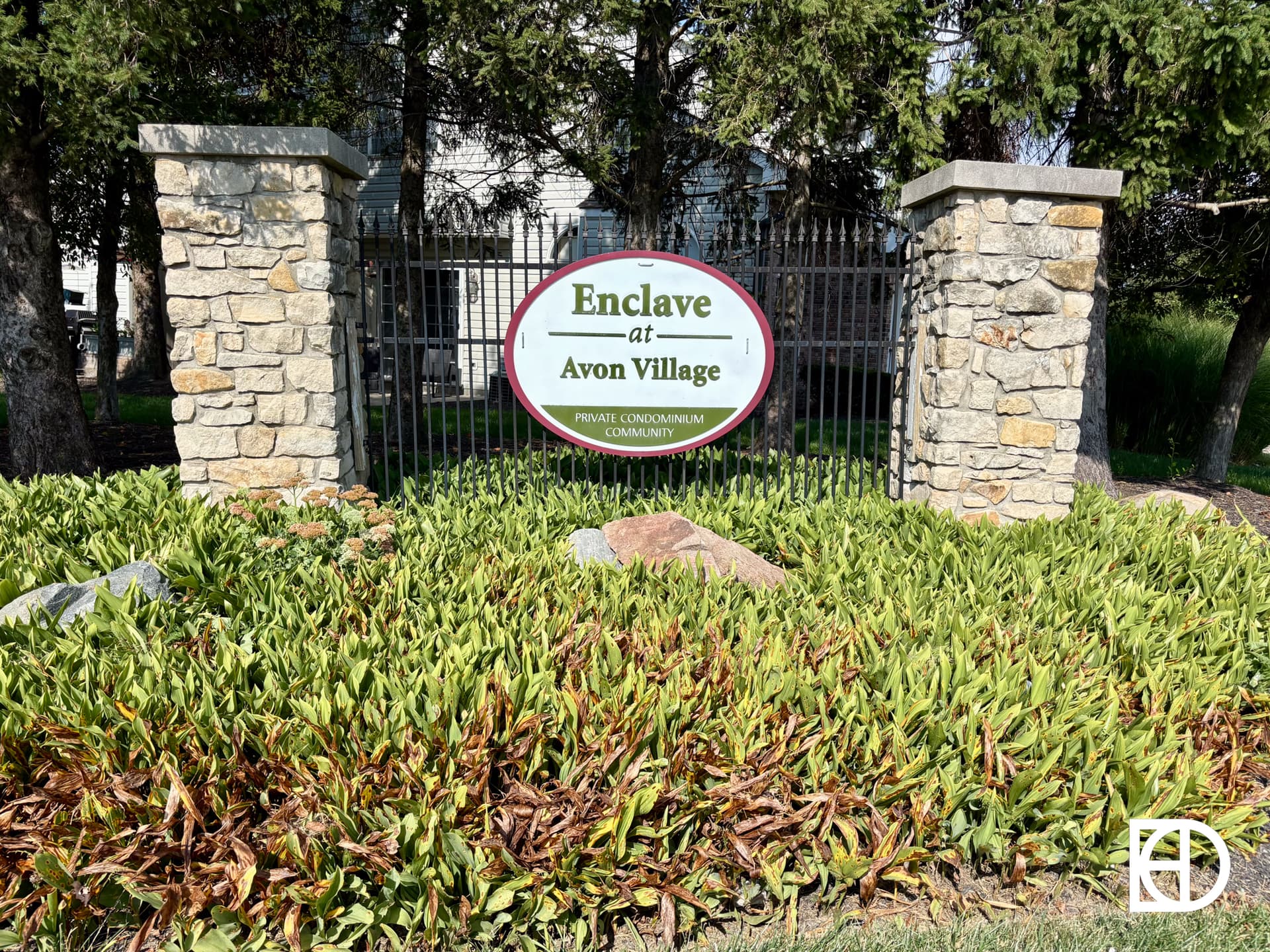 A stone sign with “Enclave at Avon Village” stands behind green foliage and in front of a metal fence, framed by stone pillars and surrounded by trees.