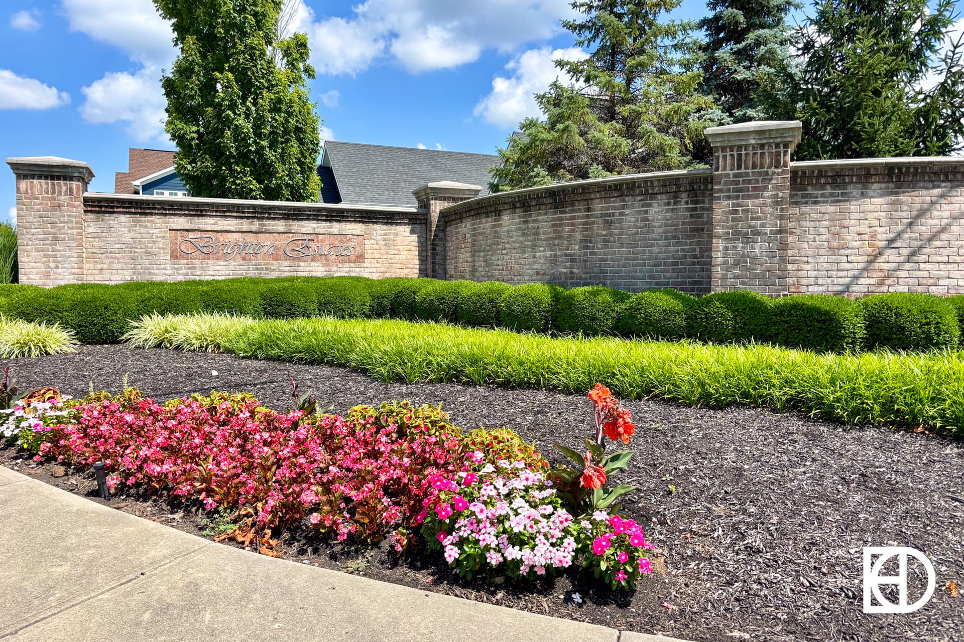 A flower bed with pink and red blooms lines a sidewalk in front of a brick wall that says Barrington Estates. Green shrubs and trees are visible behind the wall under a blue sky with scattered clouds.