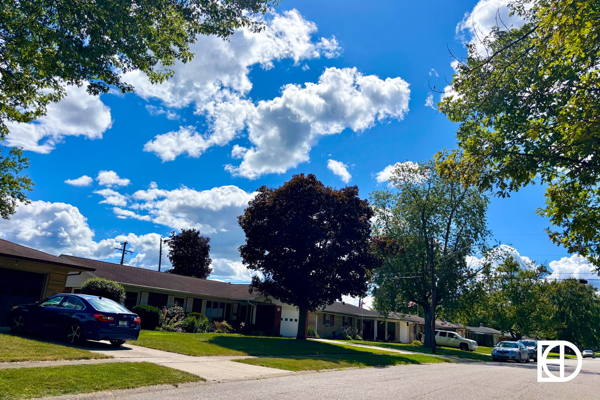 A suburban street with single-story houses, green lawns, and parked cars under a bright blue sky filled with fluffy white clouds. Trees with green and dark red leaves line the street.