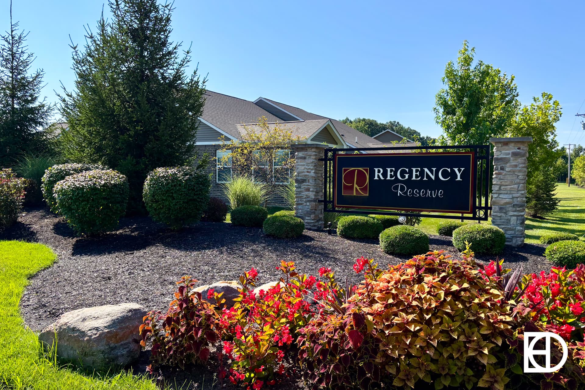 A landscaped entrance featuring a sign that reads Regency Reserve in front of a residential building, surrounded by bushes, flowers, and trees on a sunny day.