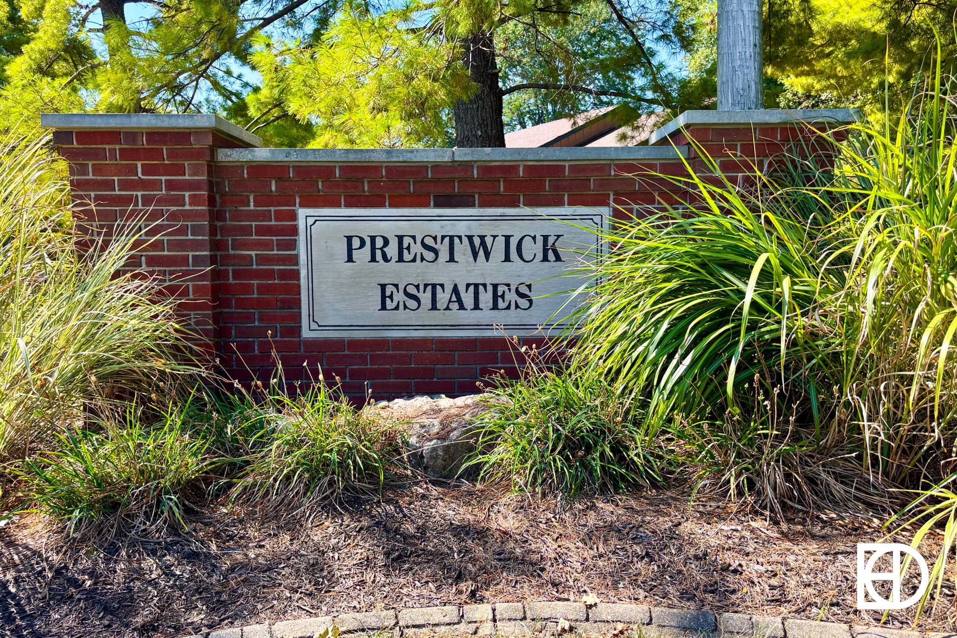 A brick entrance sign with stone plaque reading PRESTWICK ESTATES, surrounded by green plants and trees.