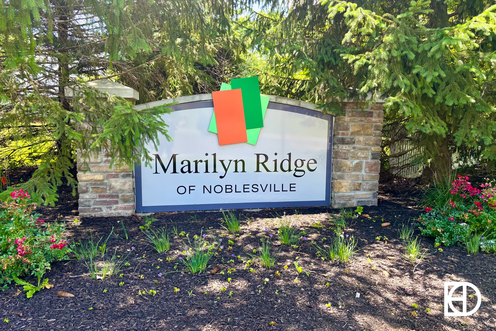 A stone sign reads Marilyn Ridge of Noblesville surrounded by green plants and red flowers. Two colored rectangles are taped to the top of the sign, and trees are visible in the background.