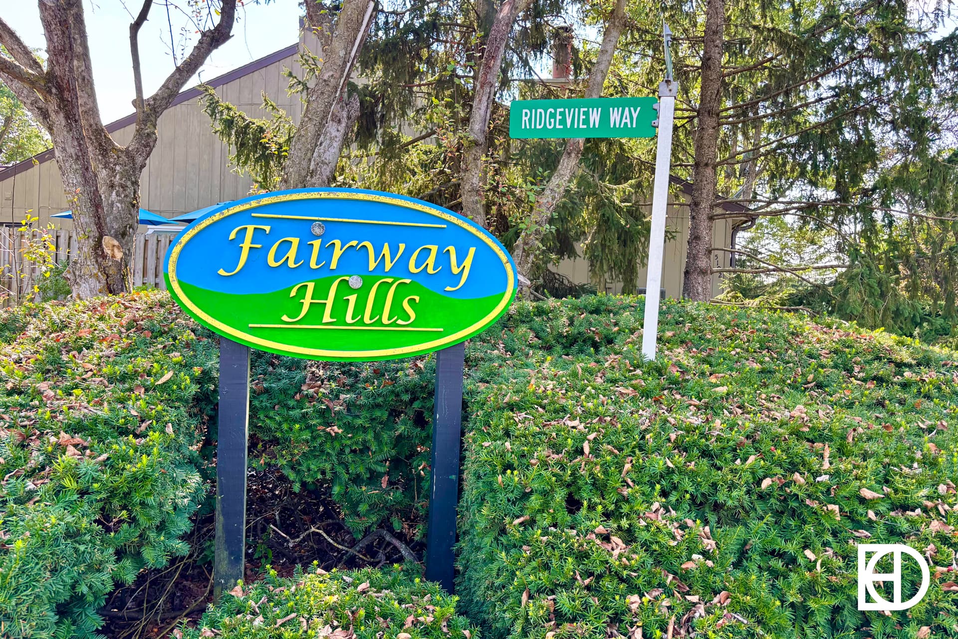 A colorful Fairway Hills sign stands among green bushes, with a Ridgeview Way street sign visible behind it and trees in the background.